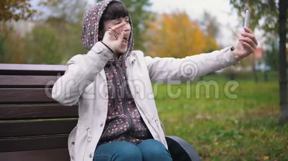 一位身穿连帽衫的年轻女子在秋季公园使用智能手机视频的预览图