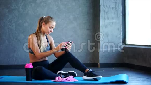 健身房里有智能手机的女人她坐在垫子上用智能手机视频的预览图