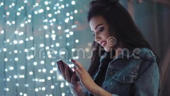 美丽的年轻女子穿着时尚的牛仔裤夹克上网在明亮的灯光下微笑夜间城市灯光视频的预览图
