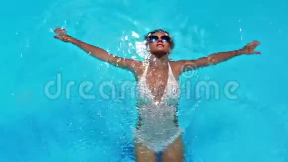 穿着白色比基尼和太阳镜的漂亮年轻女子在游泳池清澈的水中游泳视频的预览图