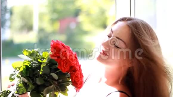 一个年轻的女人在一扇开着的窗户拿着一束红玫瑰一个美丽的女孩喜欢花香视频的预览图