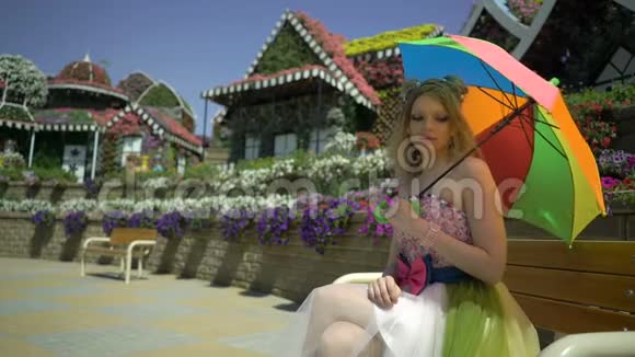 金发女郎带着彩色彩虹伞坐在花园的长凳上有趣的快乐梦中可爱的女孩视频的预览图