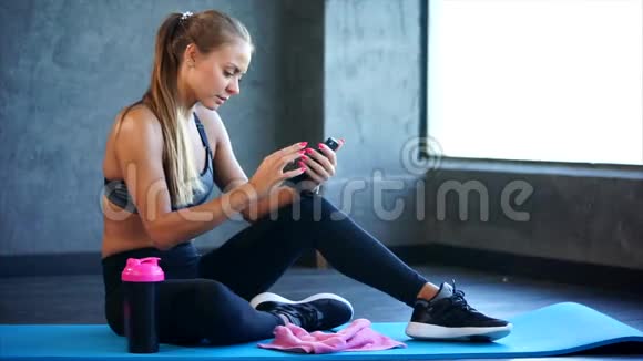 健身房里有智能手机的女人她坐在垫子上用智能手机视频的预览图