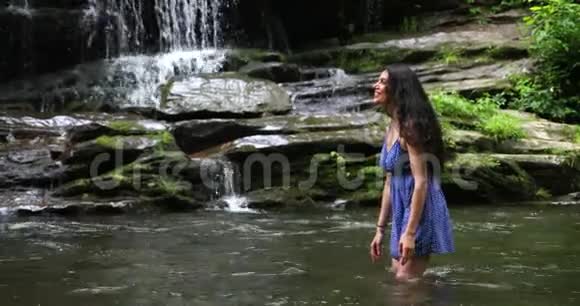 年轻女子站在瀑布附近的河里溅水女人微笑着深深地呼吸着视频的预览图