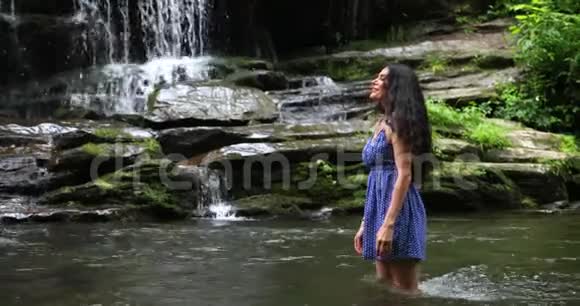 年轻的女人站在瀑布附近的河里看着落水的水女人微笑着深呼吸视频的预览图