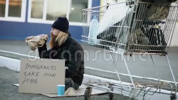 大胡子的无家可归的年轻人拿着纸板坐在购物车附近在寒冷的日子喝酒移民危机视频的预览图