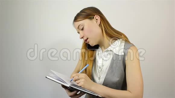 一个漂亮的年轻女孩用手机说话并在笔记本上写笔记视频的预览图