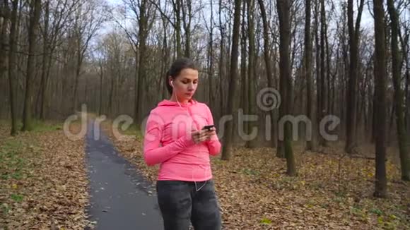 一位戴着耳机和智能手机的女士选择音乐并在日落时穿过秋天的森林慢慢视频的预览图