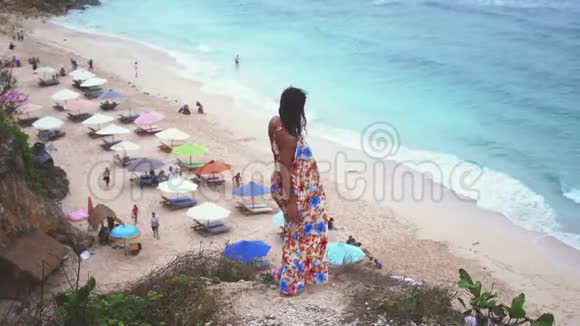多虑的年轻黑发女人穿着长裙站在海边的岩石上在刮风的天气里慢动作3840x2160视频的预览图
