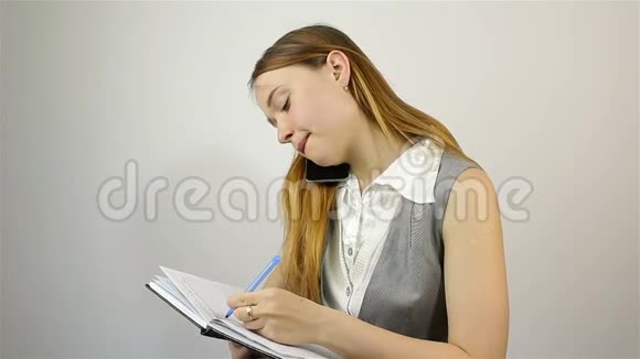 一个漂亮的年轻女孩用手机说话并在笔记本上写笔记视频的预览图