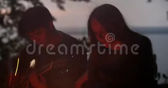 年轻的情侣坐在篝火旁在森林里弹吉他视频的预览图