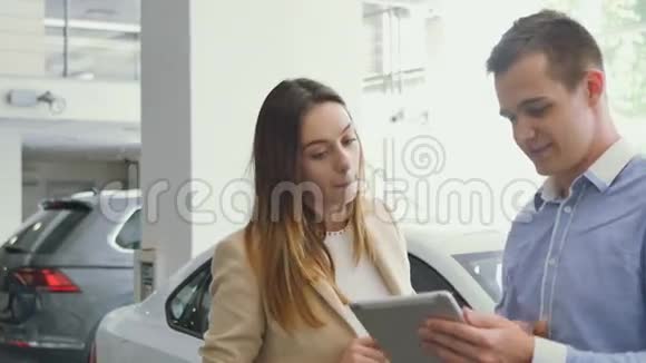 年轻人和漂亮女孩在汽车陈列室里用平板电脑视频的预览图