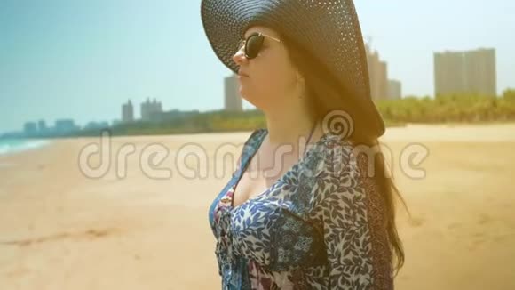 海滩上穿着蓝色连衣裙的女孩看着海洋摘下蓝色帽子近距离射击慢动作视频的预览图