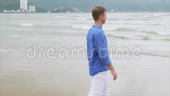 年轻微笑的金发男子穿着蓝色衬衫和白色裤子在大石头附近的海滩上散步视频的预览图