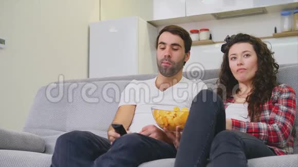 微笑的妻子和男人坐在沙发上在床上吃薯片的时候看电视穿着黑色牛仔裤视频的预览图