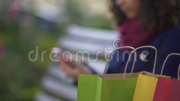 年轻女性在户外坐在长凳上在智能手机上购物视频的预览图