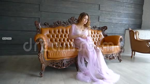 美丽迷人的孕妇棕色头发坐在沙发上穿着一件淡紫色的复古连衣裙视频的预览图