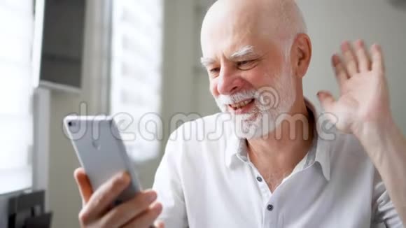 老人坐在家里拿着智能手机通过信使应用程序使用移动对话微笑着挥手致意视频的预览图