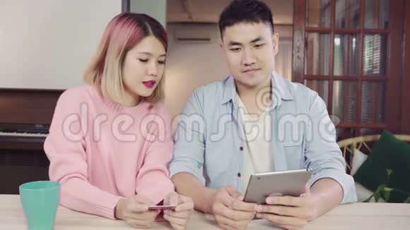 年轻的亚洲家庭夫妇使用平板电脑讨论新闻或做网上购物一起坐在办公桌上周末在家里视频的预览图
