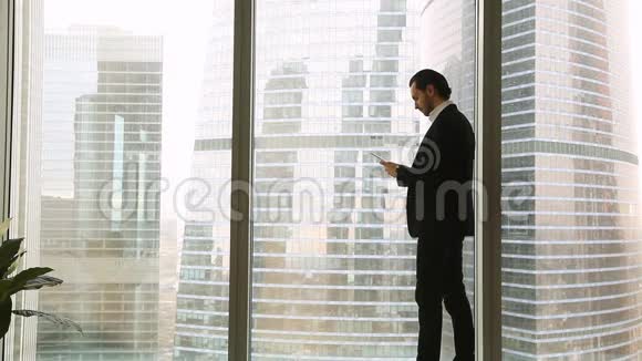 自信的年轻商人站在大窗户附近使用平板智能手机视频的预览图