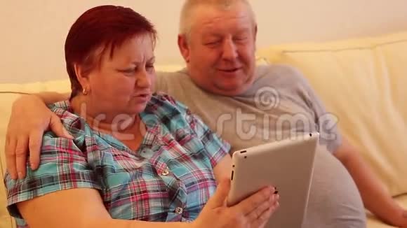 漂亮的老夫妇正在使用数码平板电脑坐在沙发上微笑视频的预览图