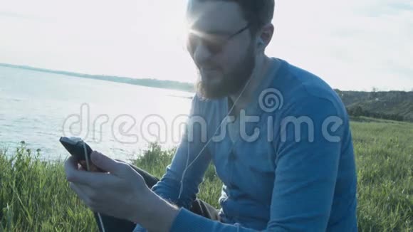 日落时分一个戴着眼镜的英俊年轻人在智能手机上写短信戴着耳机的快乐时尚男人视频的预览图