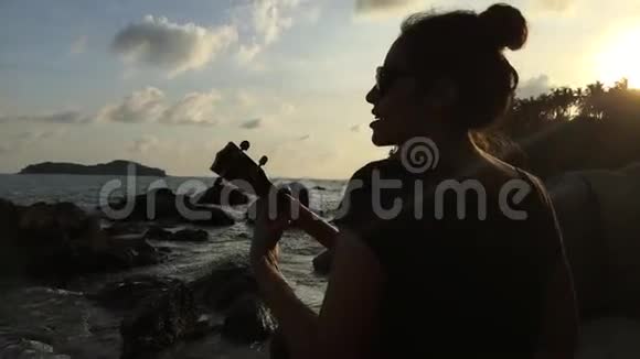 年轻的混血女孩在海滩上玩四弦琴五彩缤纷的日落高清后景银色泰国视频的预览图