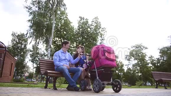 爸爸和妈妈坐在长凳上带着婴儿车在公园里家庭假日在夏天休息日视频的预览图