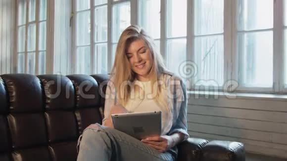 一位美丽的年轻女子正在用平板电脑翻阅一页快乐的金发女郎坐在沙发上拿着平板电脑视频的预览图