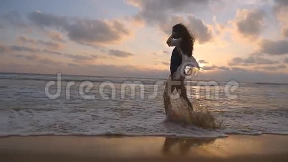 日落时穿着泳衣和衬衫在海滩散步的快乐女人海浪拍打着雌性的脚青年视频的预览图