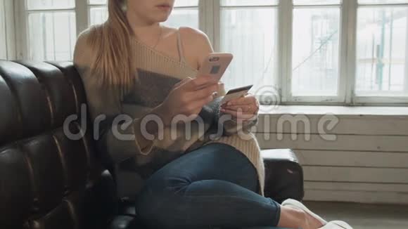 漂亮的年轻女人用银行卡通过智能手机购买一个金发女郎坐在家里的椅子上视频的预览图