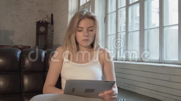 沙发上一个漂亮的年轻金发女郎的肖像带着一张体贴的平板电脑在一个大房间里有一个钟和一个旧的视频的预览图