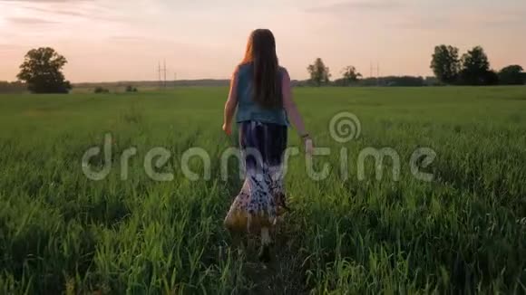 年轻的女孩带着微笑直发穿着一件长裙沿着绿色的田野奔跑夕阳西下慢动作视频的预览图