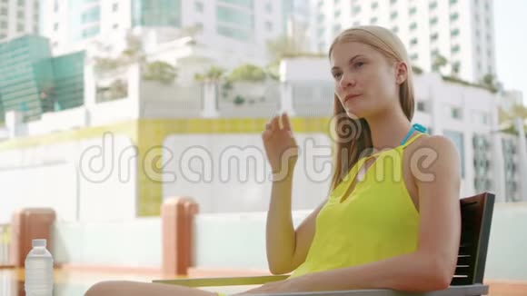 一位穿着黄色连衣裙坐在游泳池旁的椅子上扭着头发的美丽梦幻的年轻女子视频的预览图