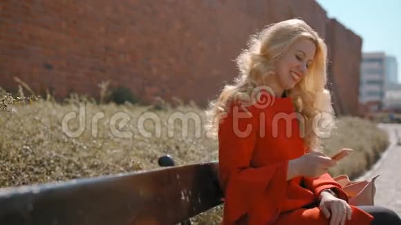 美丽的金发女人坐在长凳上用她的移动智能手机应用程序视频的预览图