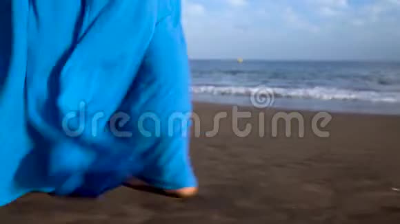 一个穿着漂亮蓝色裙子的女人在黑色火山海滩上行走的腿视频的预览图