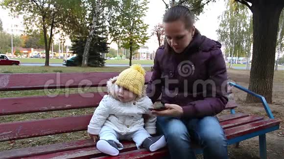 妈妈带着孩子坐在公园的长凳上妈妈给你的孩子看一部智能手机视频的预览图
