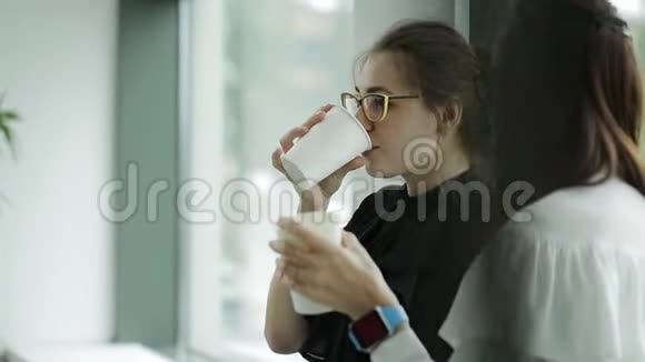 办公室窗口附近的两个女商人在一天结束时喝着咖啡和闲聊视频的预览图