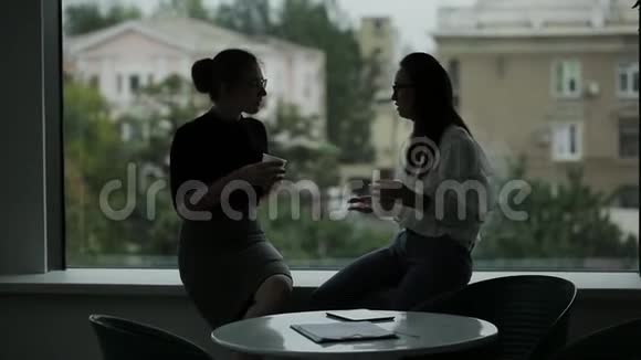 办公室窗口附近的两个女商人在一天结束时喝着咖啡和闲聊视频的预览图