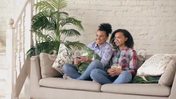 两个混血卷发女孩朋友坐在沙发上看喜剧电影吃爆米花有趣的姐妹视频的预览图