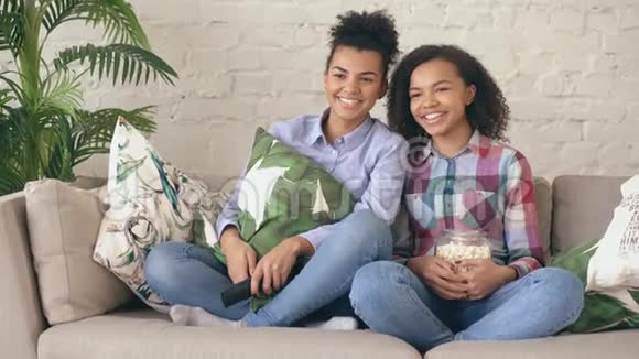 两个混血卷发女孩朋友坐在沙发上看喜剧电影吃爆米花有趣的姐妹视频的预览图