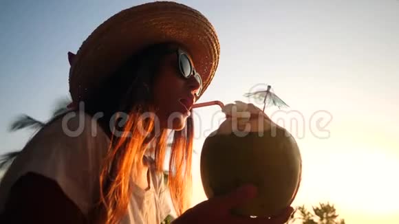 迷人的年轻旅游少女在美丽的日落海滩喝新鲜的泰国椰子水可可4K视频的预览图
