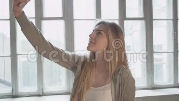 赛尔夫年轻漂亮的金发女郎拿着一部手机对着一扇大窗户送一个空气吻在一个视频的预览图