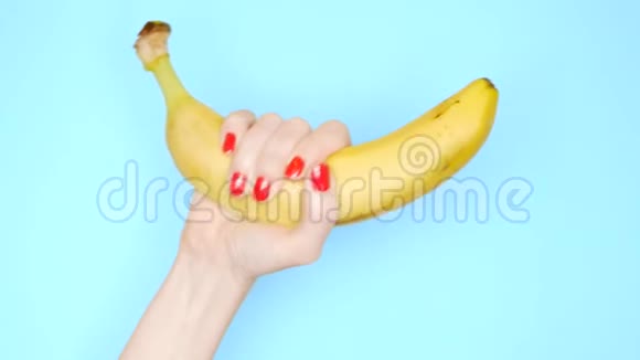 有红色指甲的女性手在蓝色背景上拿着一根黄色的香蕉视频的预览图