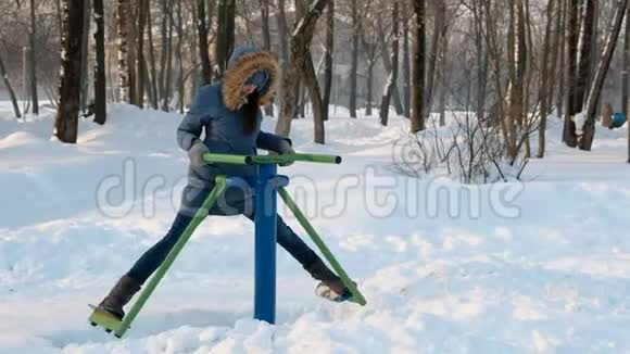 穿着蓝羽绒服带头罩的女人在冬季公园前景的模拟器上做腿部运动视频的预览图