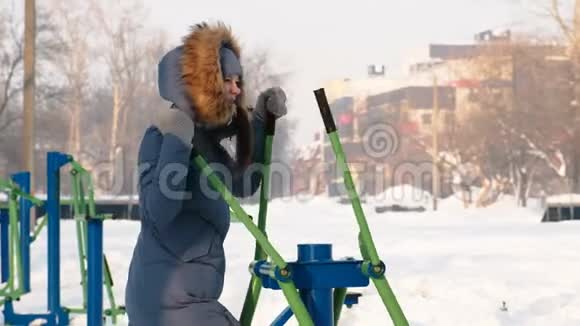 身穿蓝羽绒服头罩的女人在城市的冬季公园里从事滑雪模拟器视频的预览图