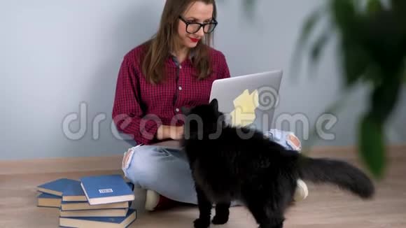 漂亮的女孩用笔记本电脑抚摸一只猫坐在家里的地板上视频的预览图