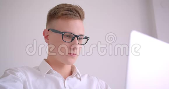 年轻英俊的白种人商人戴着眼镜用笔记本电脑看着镜头微笑的特写肖像视频的预览图