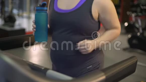 超重妇女在跑步机上运动从瓶子里喝水饮食视频的预览图