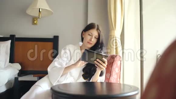 穿着浴袍的年轻女商人使用平板电脑坐在酒店房间的椅子上视频的预览图